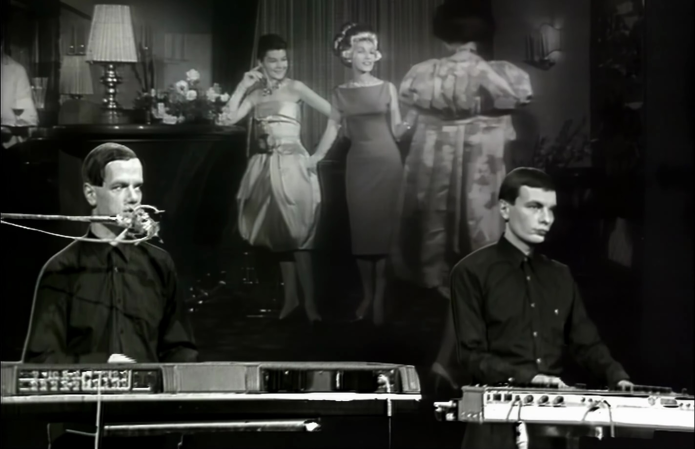 A shot of the video for the Kraftwerk song Das Model with the band standing behind synthesizers in front of a film of a 1950s model show.